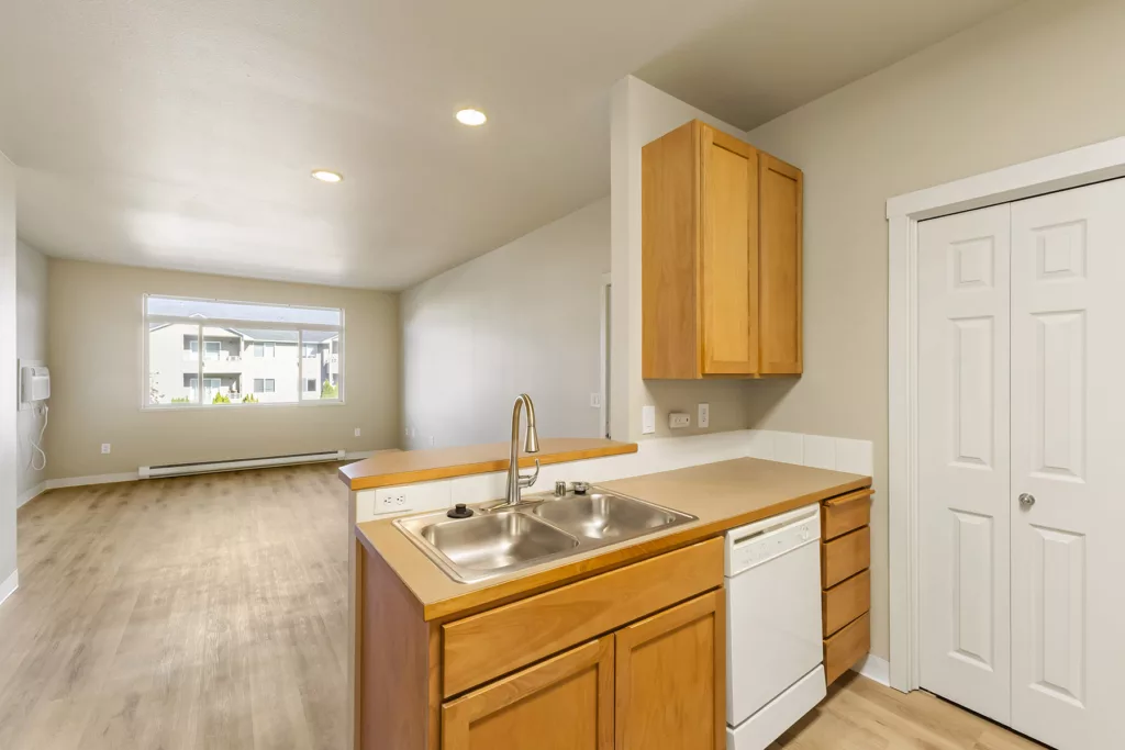 Open-plan kitchen with double sink, wood cabinets, dishwasher, and a large window looking into the living area