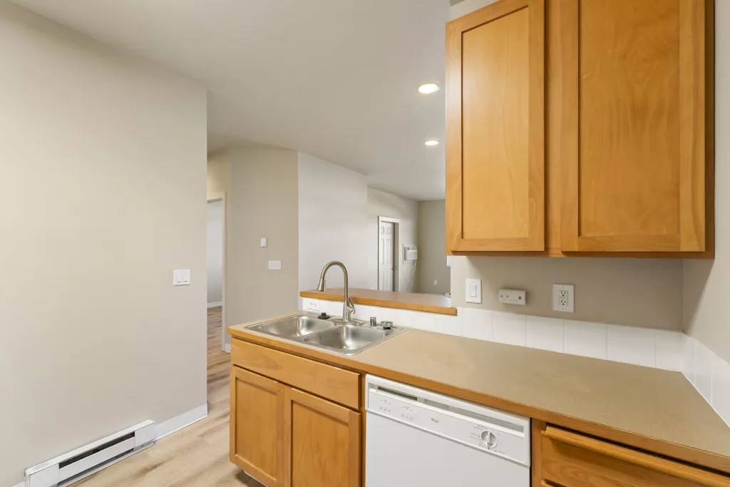 Kitchen counter with double sink, faucet, dishwasher, and wooden cabinets, overlooking an open hallway.