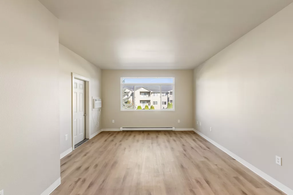 Empty living room with light walls, wood flooring, a large window, and a door with a wall-mounted heater nearby