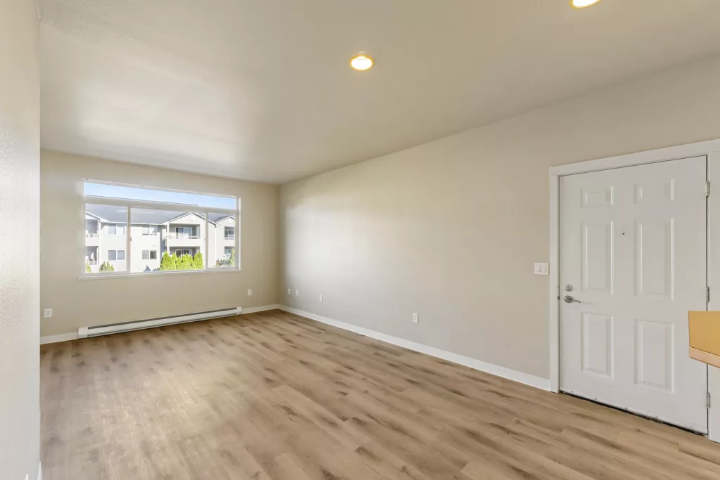 Empty living room with wood-look flooring, beige walls, recessed lights, large window, and white front door