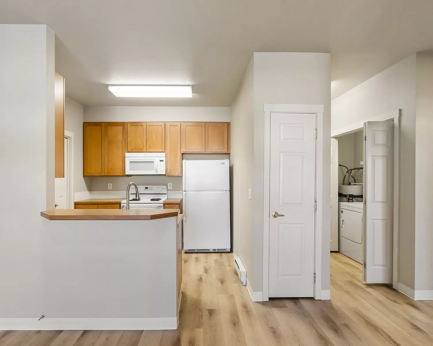 Bright apartment kitchen with wood cabinets, white fridge and microwave, breakfast bar, and open laundry closet.