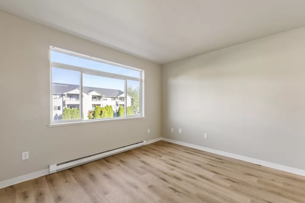 Empty room with light wood floors and a large window overlooking nearby apartment buildings and trees