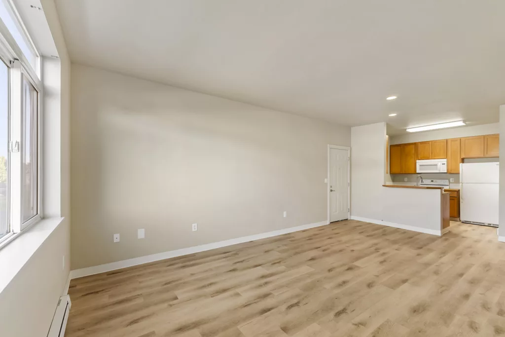 Empty living room with large window, light wood floors, and open kitchen with wood cabinets and white appliances