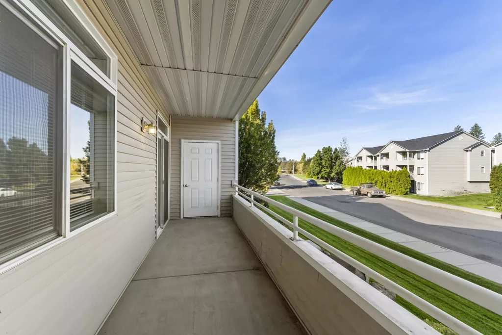 Covered apartment balcony with railing, sliding door and storage door, overlooking a quiet street and nearby buildings.