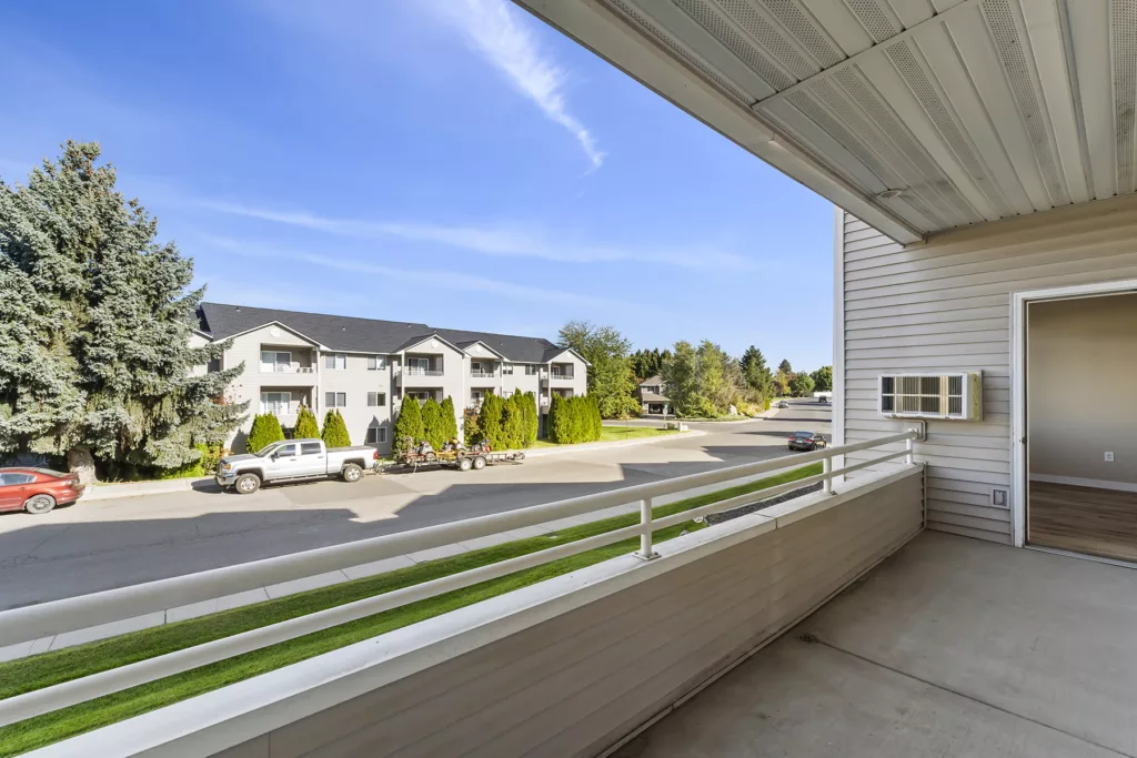 Balcony view overlooking a quiet street, apartment buildings, parked vehicles, and trees under a clear blue sky.