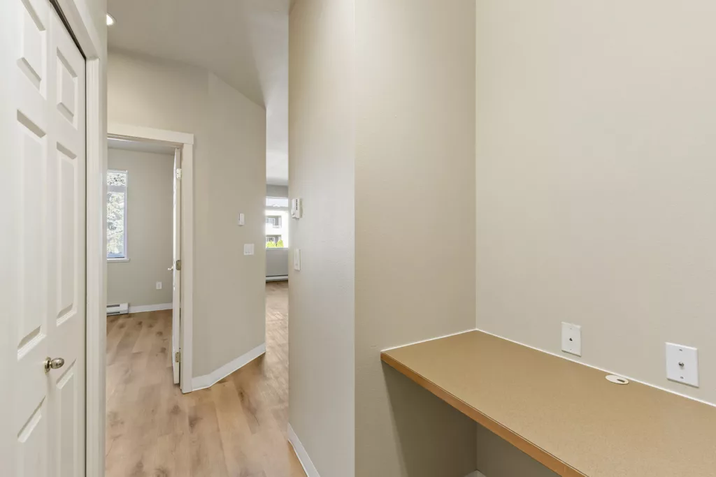 Hallway with light wood floors, open doorway to a room, and a built-in desk nook with outlets on the wall