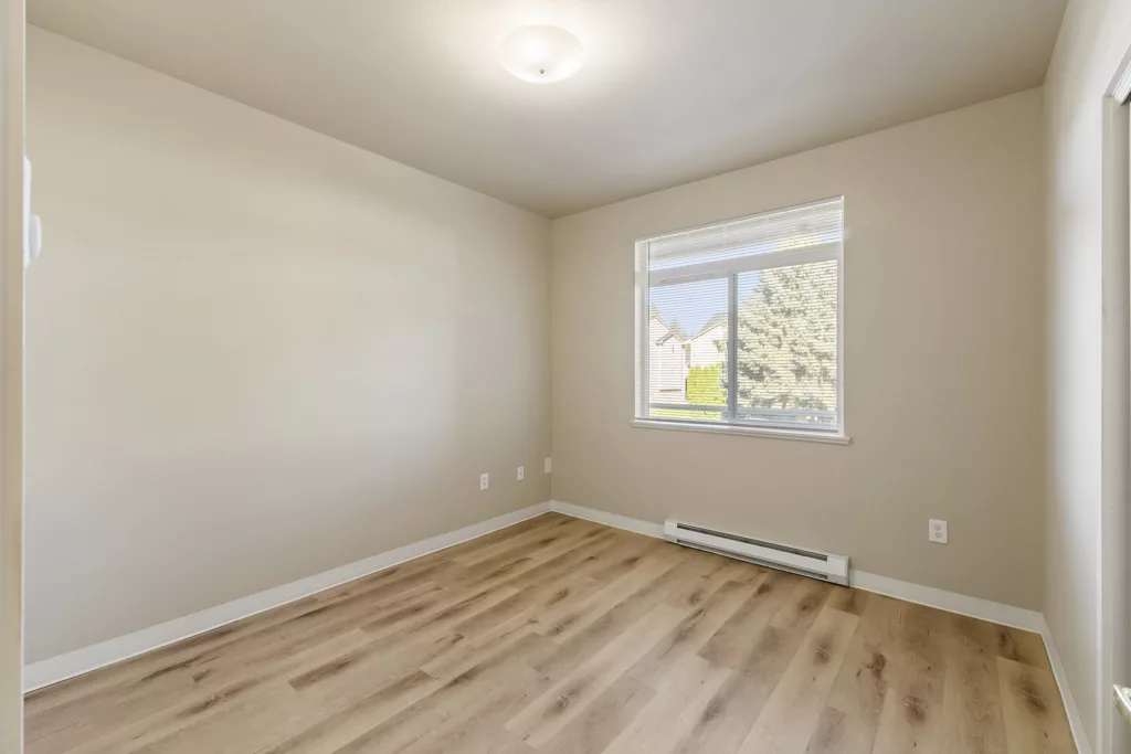 Empty beige room with wood-look flooring, ceiling light, and a window with blinds above a baseboard heater.