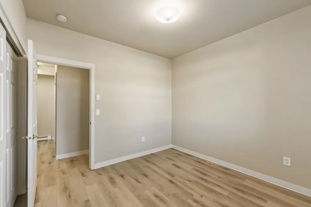 Empty beige room with light wood floors, ceiling light, and open doorway to a hallway or closet