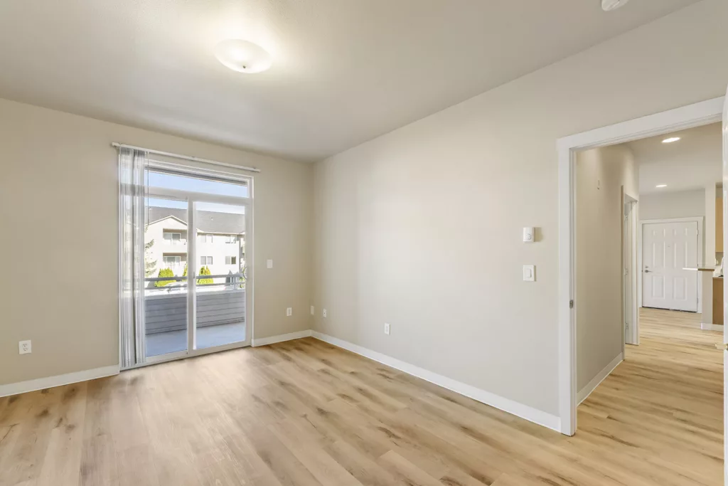 Empty room with wood floors, beige walls, and sliding glass door to a balcony, plus hallway entry on the right