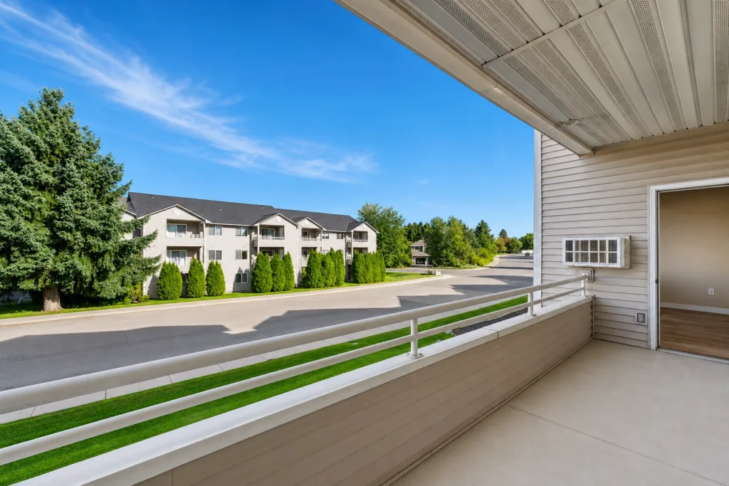 Balcony view overlooking a quiet street with apartment buildings, trees, and a bright blue sky.