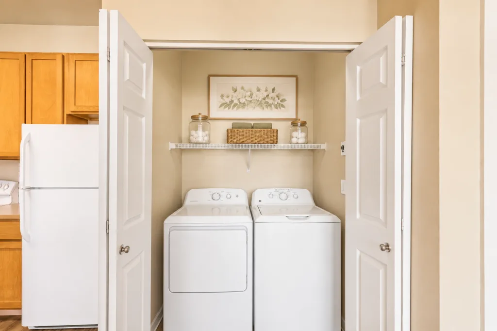 Washer and dryer in a closet with open double doors, shelf above with jars and basket, next to a kitchen fridge