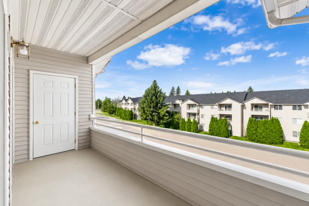 Covered apartment balcony with white door and railing overlooking neighboring buildings, trees, and a blue sky