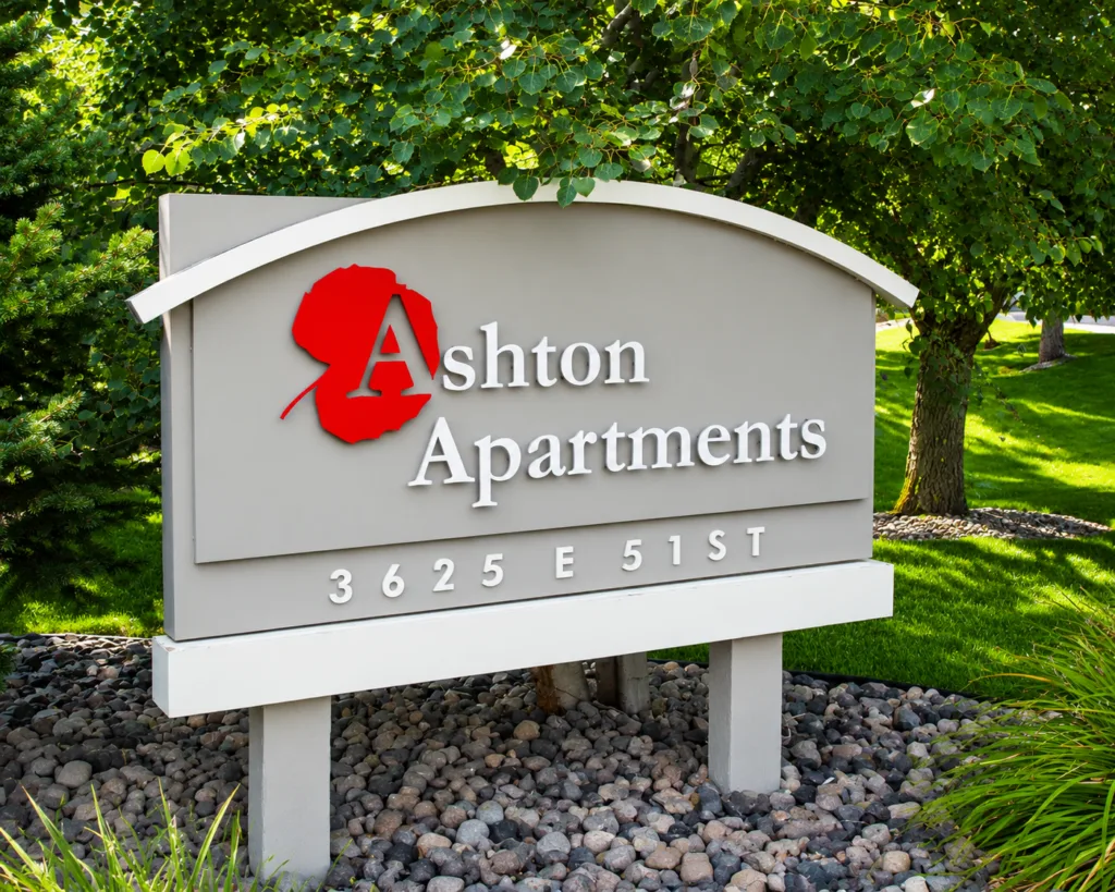 Ashton Apartments entrance sign with red logo, showing address 3625 E 51st amid trees and landscaping