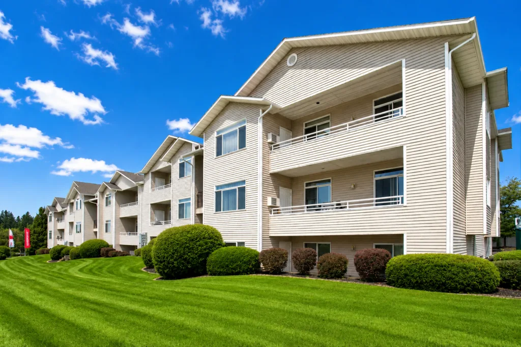 Row of beige apartment buildings with balconies beside a manicured green lawn under a bright blue sky
