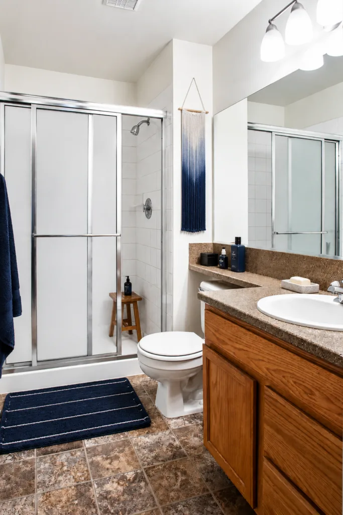 Bright bathroom with sliding glass shower, toilet, wood vanity with sink, large mirror, and navy bath mat