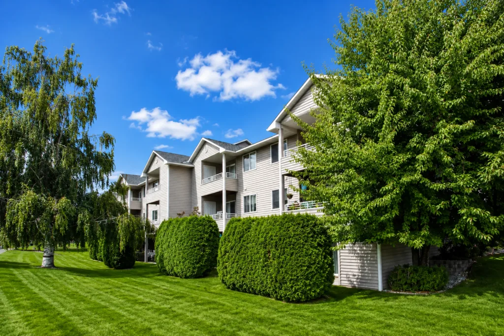 Apartment building with balconies beside a manicured lawn, shrubs, and leafy trees under a bright blue sky.