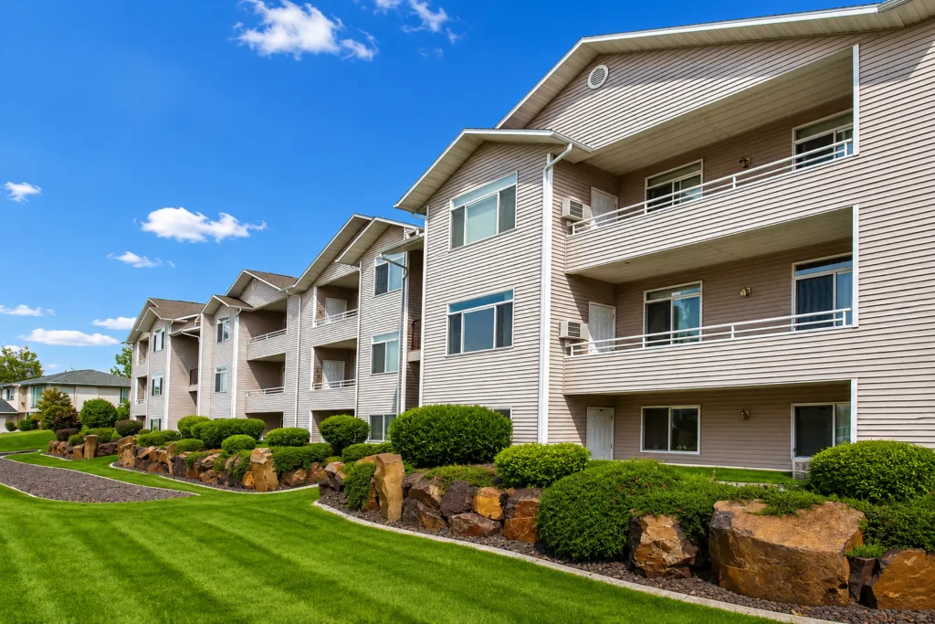 Exterior of a three-story apartment building with balconies, landscaped lawn, shrubs, and decorative rocks under blue sky