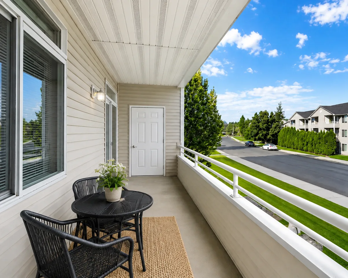 Apartment balcony with two chairs and a small table, overlooking a quiet suburban street and houses.